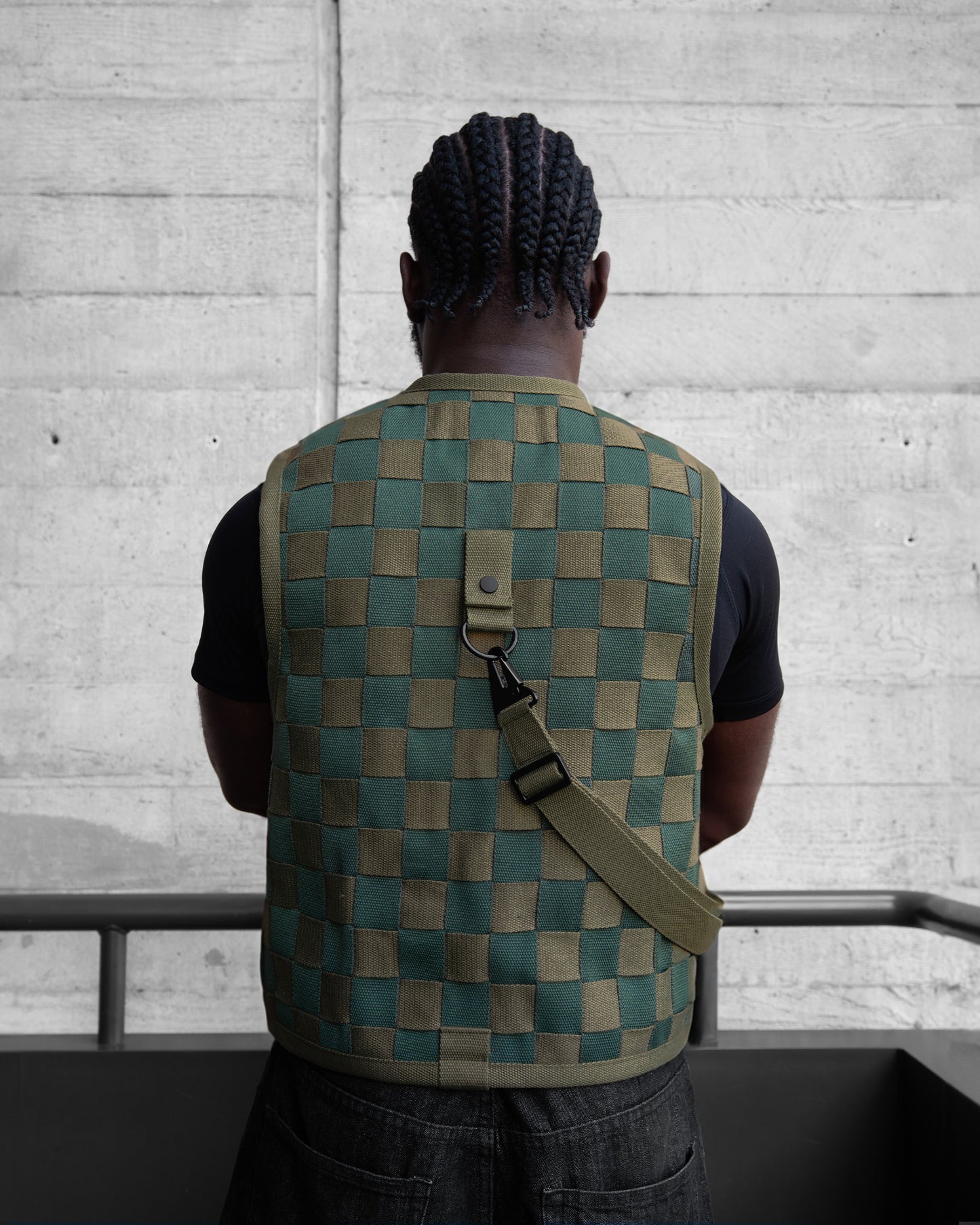 Person wearing a green checkered vest with a bag over their shoulder against a concrete wall.