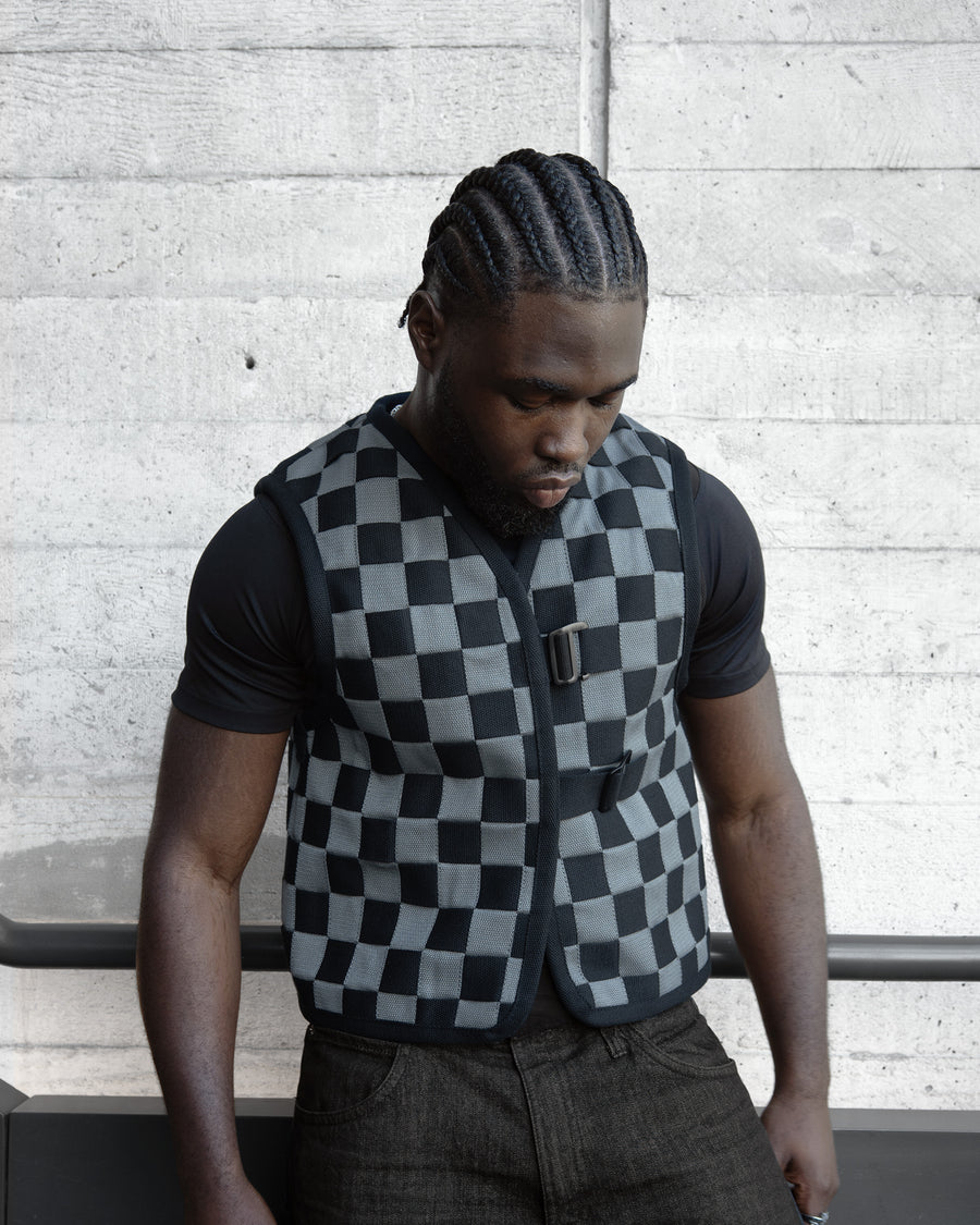Man wearing a checkered vest against a concrete wall.