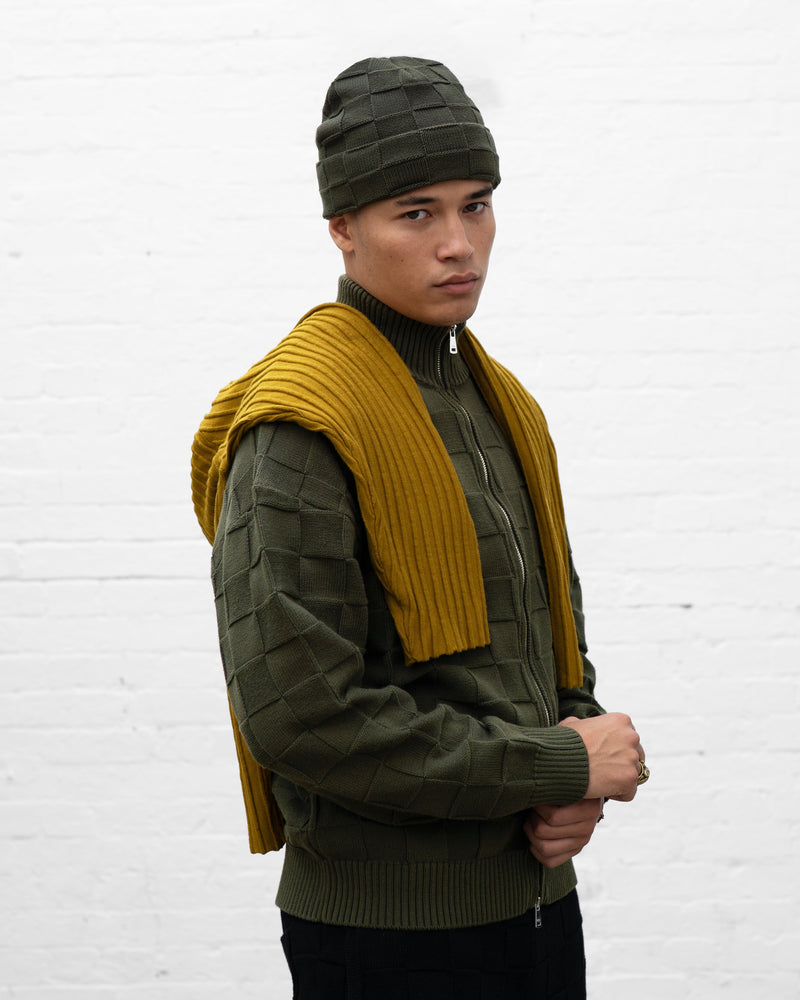 A man wearing a dark olive green textured knit beanie, a matching zip-up sweater vest, and a mustard yellow ribbed knit sweater draped over his shoulders, standing against a white brick wall.