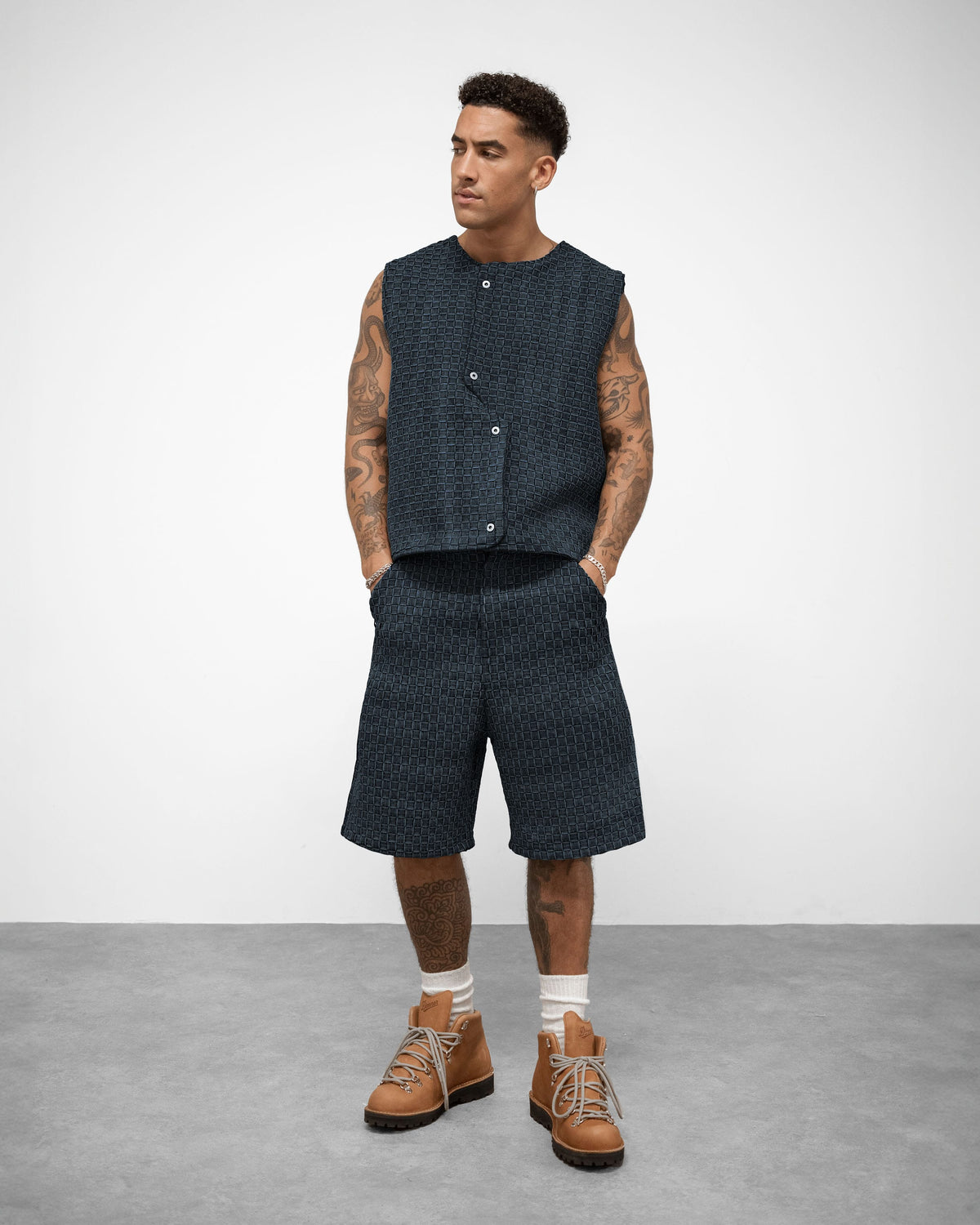 Man wearing BRYDER woven denim dark sleeveless vest, shorts, and brown boots on a plain background